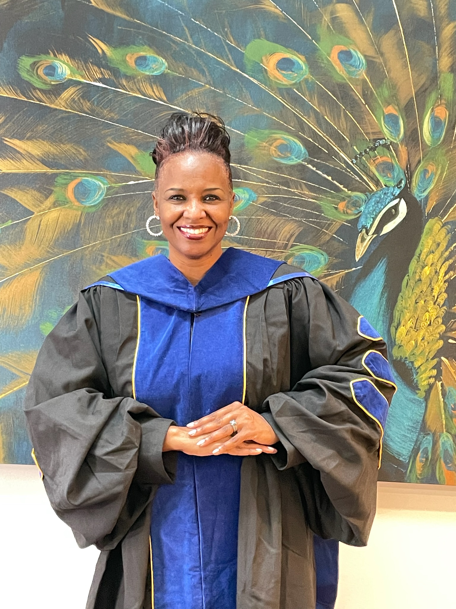 Dr. DeAnna Lynn Nix in doctoral regalia standing before a peacock backdrop